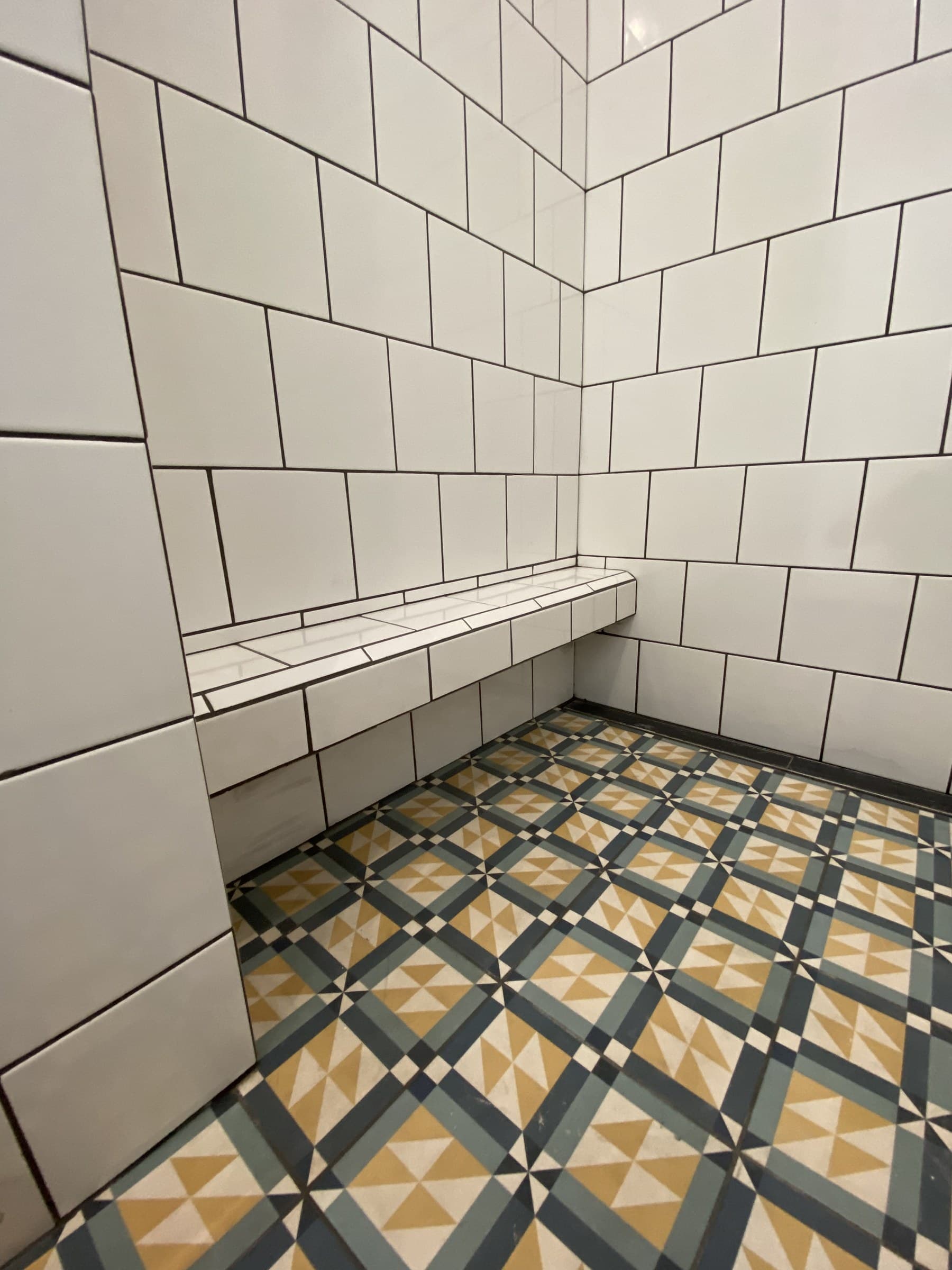 Corner of a white square-tiled shower with a tiled bench and a patterned blue, mustard and cream floor